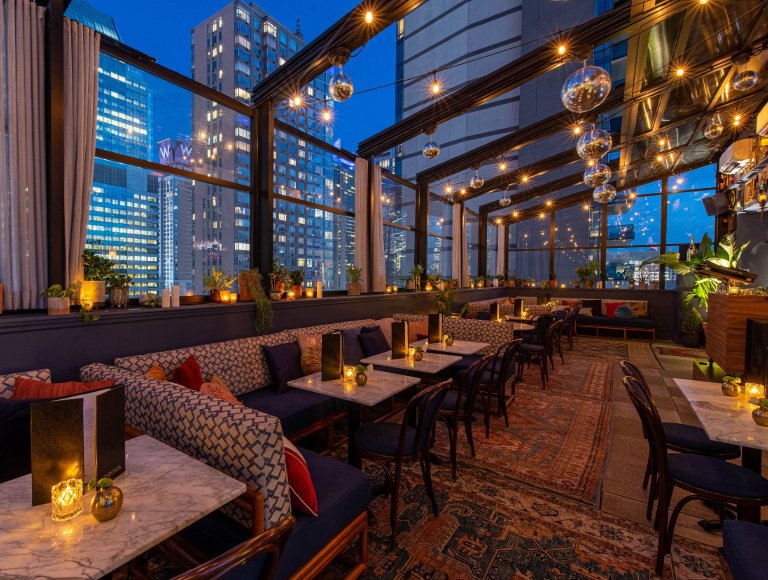 Starchild Rooftop bar at dusk with glass walls over city towers, globe string lights, and banquette seating with marble tables
